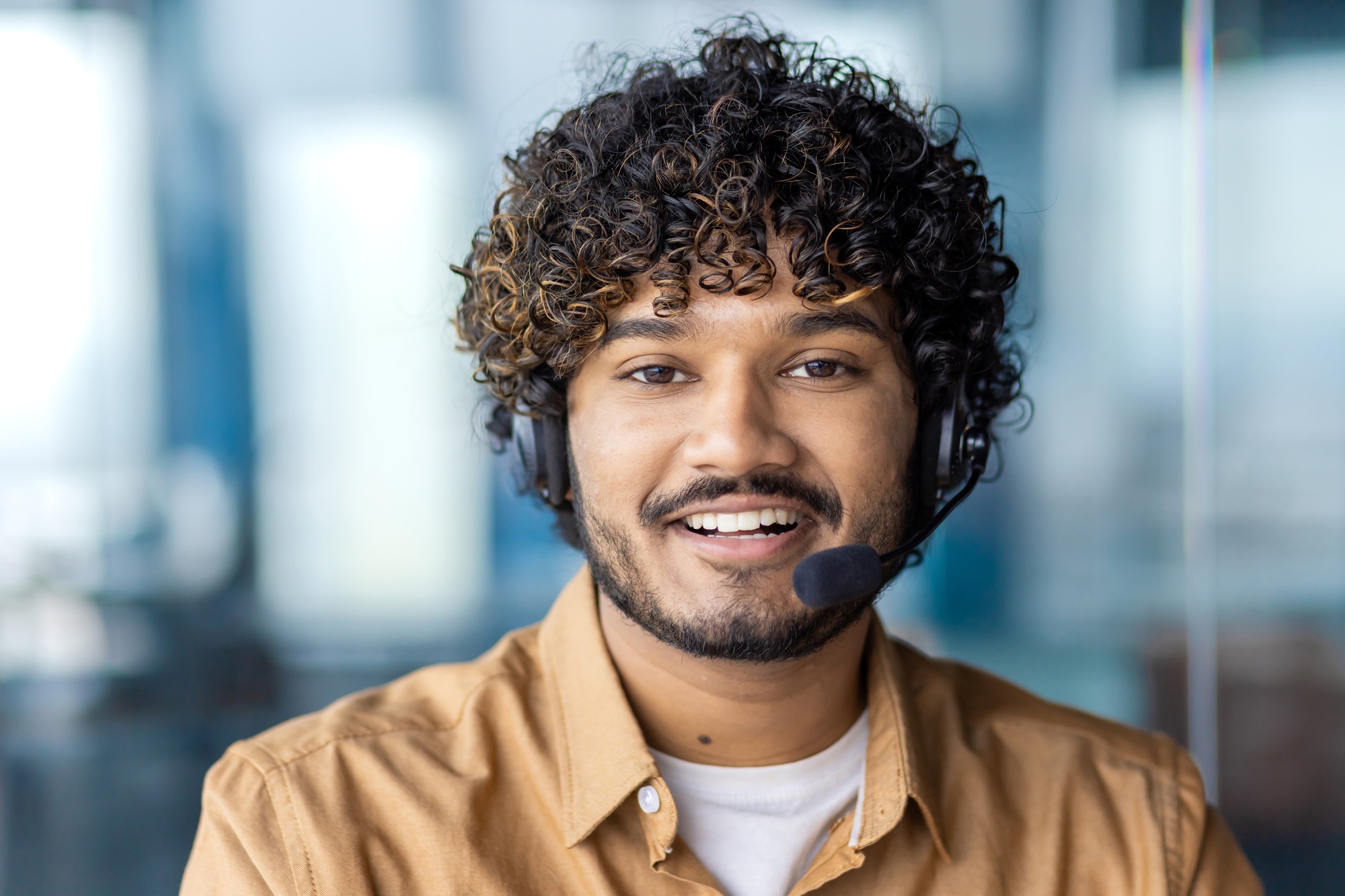 Portrait of positive indian guy wearing headset with microphone and looking at camera on blurred